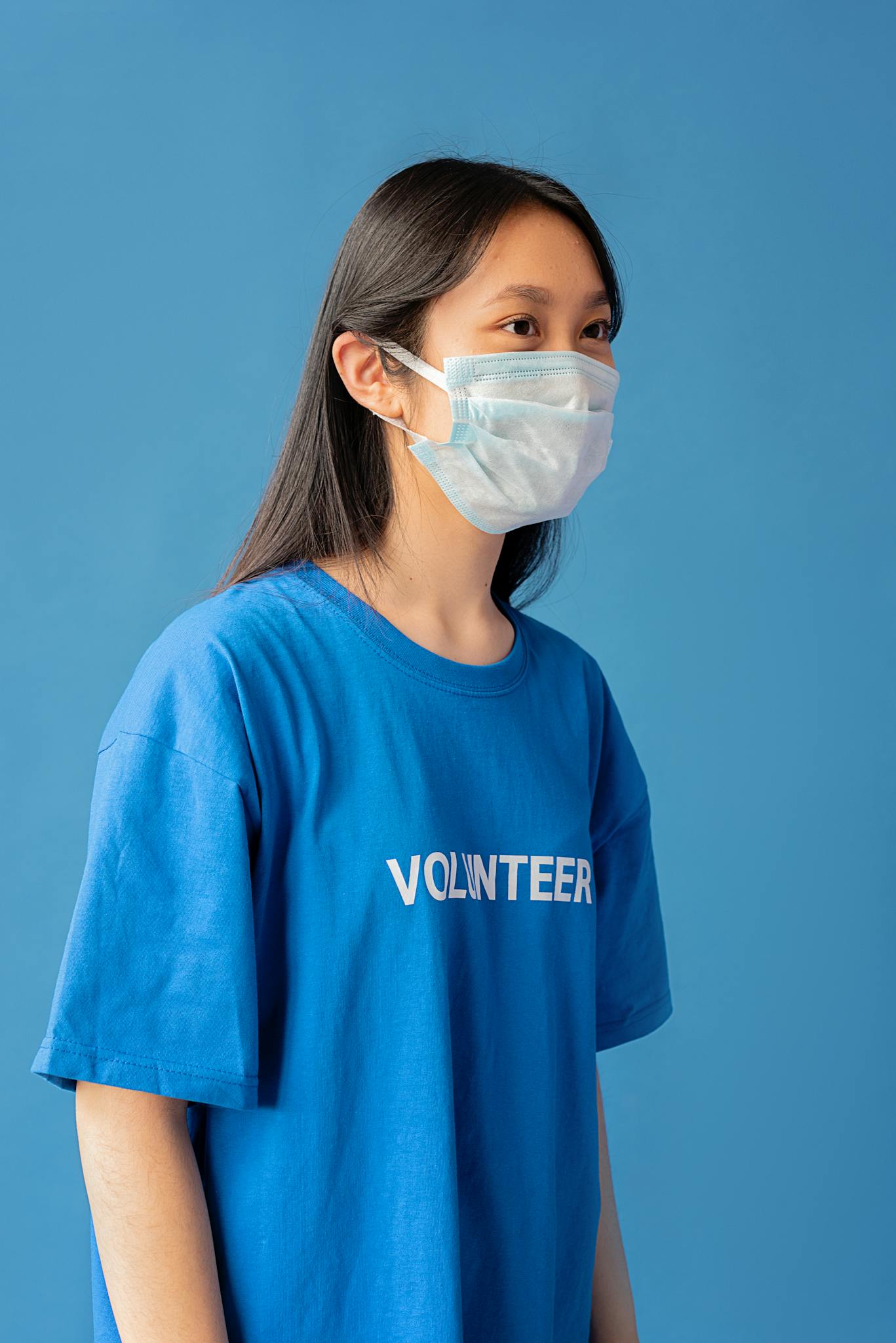Portrait of an Asian woman in a mask and blue shirt labeled 'Volunteer,' symbolizing pandemic volunteer efforts.